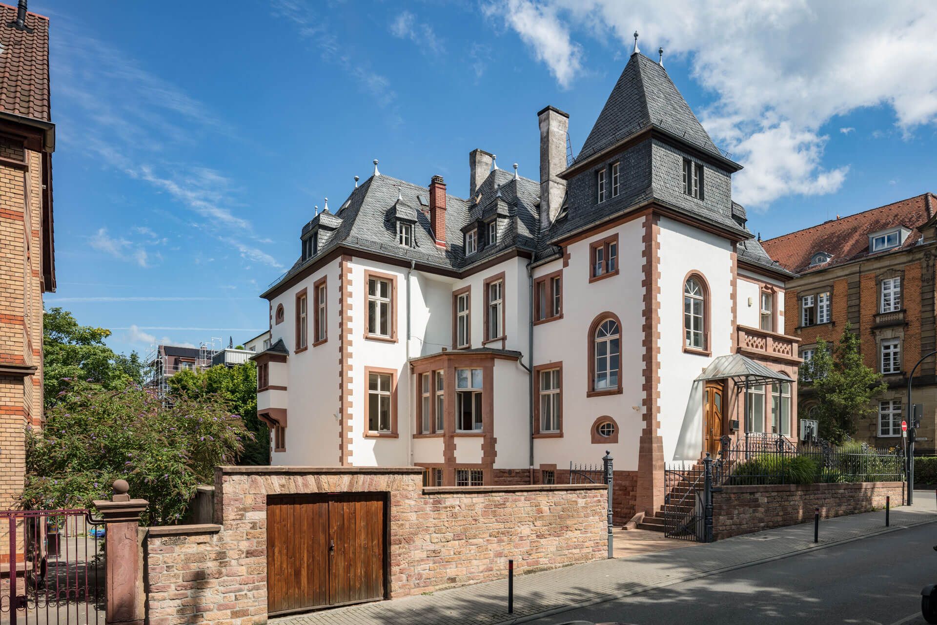 Elegantes weißes Backsteinhaus mit Schieferdach, umgeben von Grünflächen und Kopfsteinpflaster unter blauem Himmel.