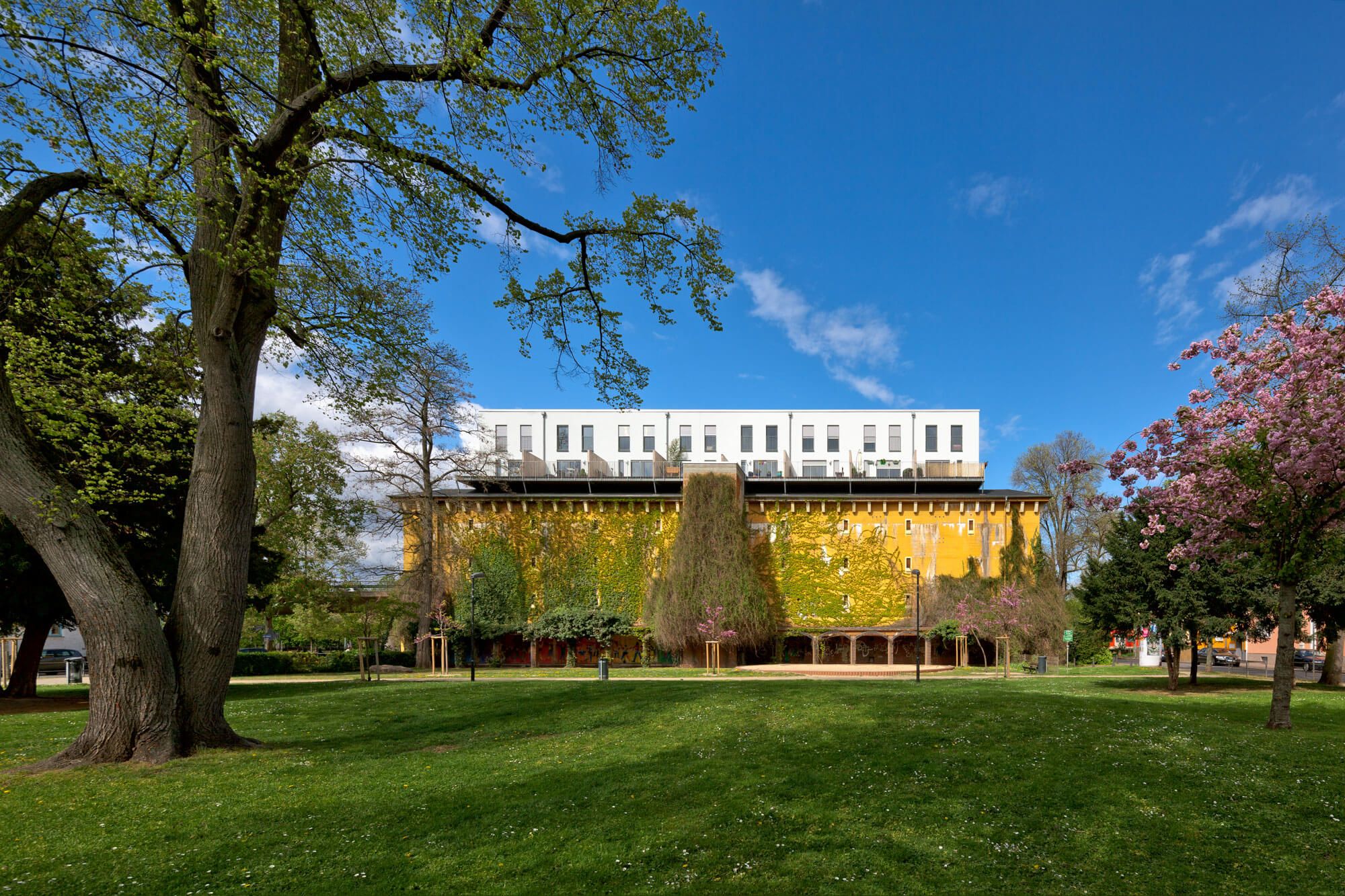 Ein leuchtend gelbes Gebäude mit grünem Laub, das von modernen weißen Wohnungen gekrönt wird, vor einem klaren blauen Himmel und einem grünen Park.