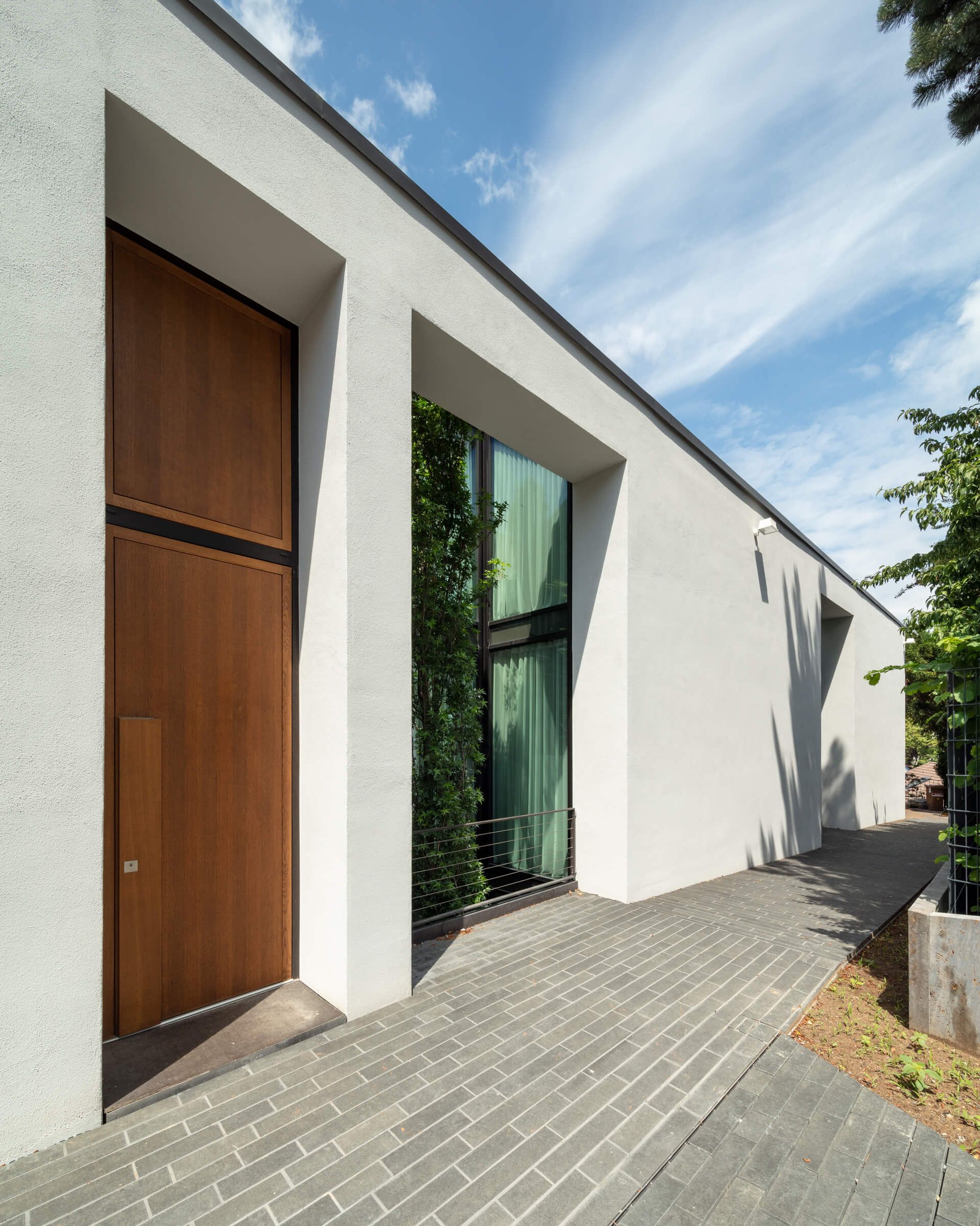 Ein modernes weißes Gebäude mit einer Holztür, großen Glasfenstern und einem gepflegten Weg unter einem blauen Himmel mit Wolken.