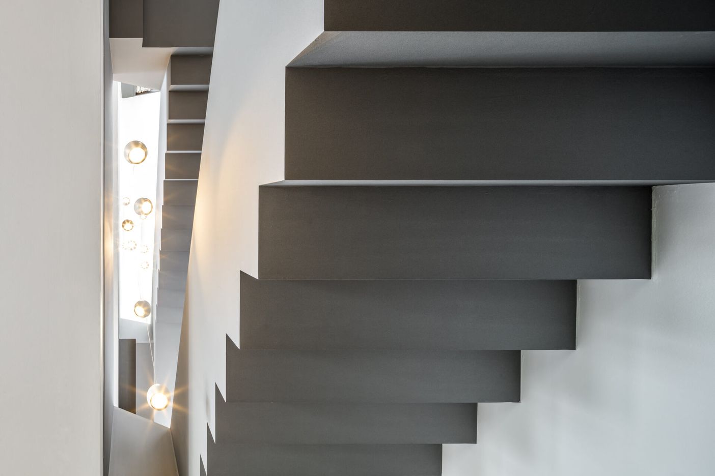 Eine Nahaufnahme moderner, freitragender schwarzer Treppenstufen neben einer beleuchteten, minimalistischen Wand und dezenten Beleuchtungskörpern.